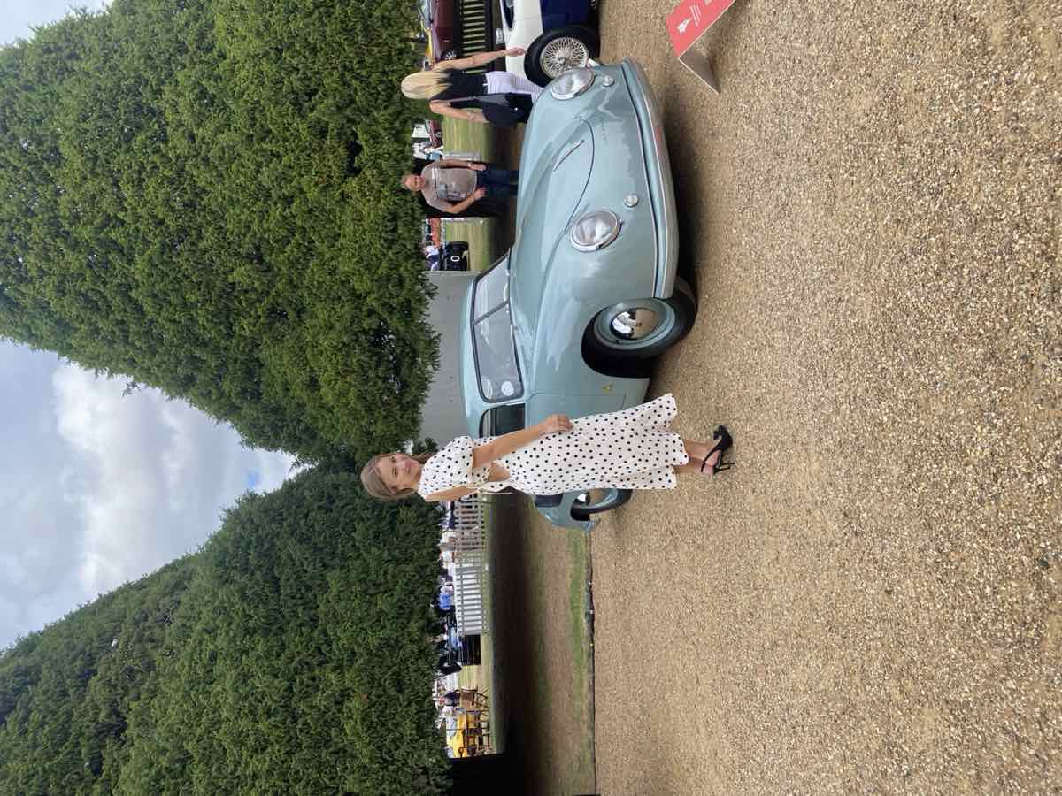 Taylor beside a pale blue vintage car between tall clipped hedges