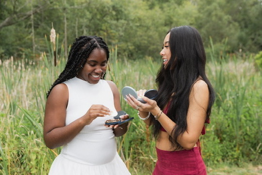 Two friends laughing and sharing a compact in front of tall meadow grasses, from the Golden Bubble shoot
