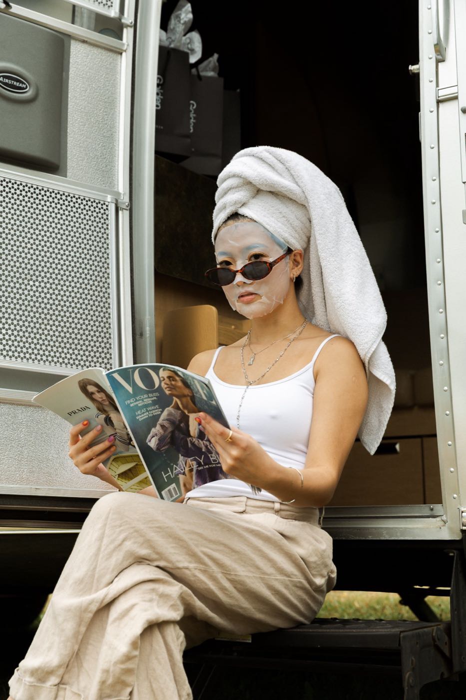 Model in a sheet mask and sunglasses reading a magazine on the step of an Airstream